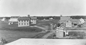 Black-and-white photo of a rural village with several wooden buildings, unpaved roads, fences, and sparse trees in the background. The scene appears quiet and historic, likely from the 19th or early 20th century.