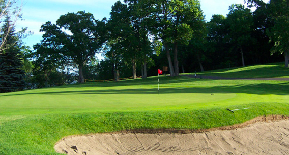 A lush golf course with a sand bunker in the foreground, a red flag on the putting green, and tall trees in the background beneath a clear blue sky.