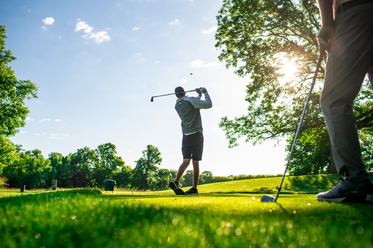 A person swings a golf club on a lush, green golf course under a bright, sunny sky, with trees and another person standing nearby in the background.