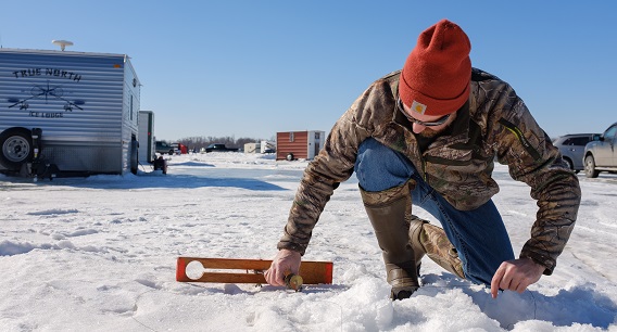 A person in a red beanie and camouflage jacket kneels on snow, using a level tool on the ice. Several trailers and vehicles are visible in the background under a clear blue sky.