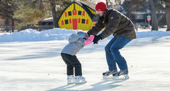 An adult and a child ice skating outdoors on a sunny winter day. The adult is helping the child balance. Both are wearing winter jackets and hats, with snow and a yellow building in the background.