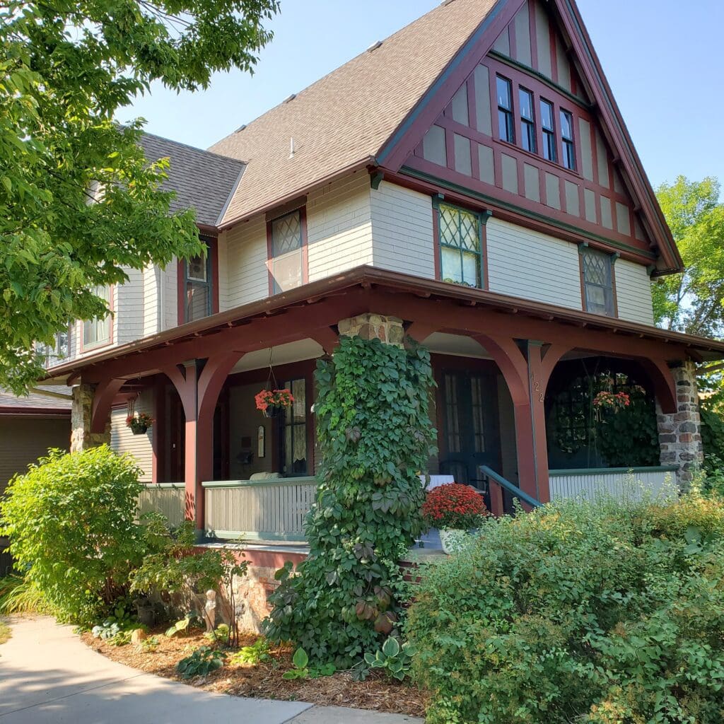 A two-story house with a sloped roof, Tudor-style detailing, and maroon trim, Cedar Rose Inn is a charming bed and breakfast Alexandria gem, featuring a vine-covered porch surrounded by lush greenery and hanging baskets of red flowers.