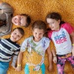 Four children lie side by side, smiling up at the camera on a bed of yellow corn kernels at Country Blossom Farm. Dressed in colorful, casual clothes, they radiate joy and playful energy in this cheerful moment.