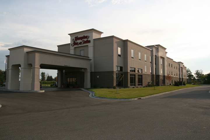 A three-story Hampton by Hilton Inn & Suites hotel building with a covered entrance, surrounded by grass and a paved driveway under a partly cloudy sky.