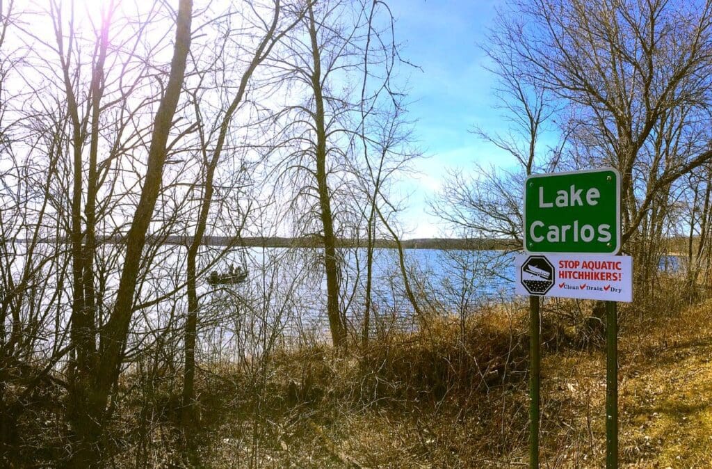 A green "Lake Carlos" sign stands near leafless trees by the water, with a boat on the lake and a warning below about stopping aquatic hitchhikers—perfect for easy spring adventures at Lake Carlos State Park under a clear, sunny sky.