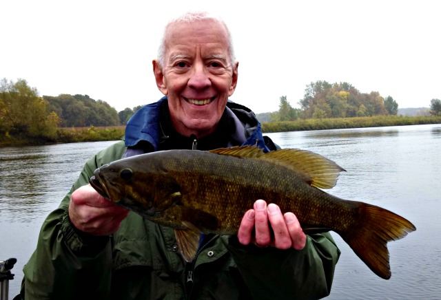 An older man in a green jacket smiles while holding a large fish in front of a river, showcasing his success with fall smallmouth bass fishing tips. Trees and cloudy skies set the scenic background.