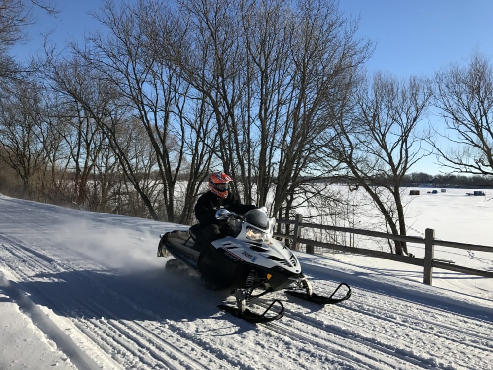 A person in winter gear rides a snowmobile along Alexandria snowmobile trails, gliding past a wooden fence with bare trees and a snowy landscape in the Alexandria area under a clear blue sky.