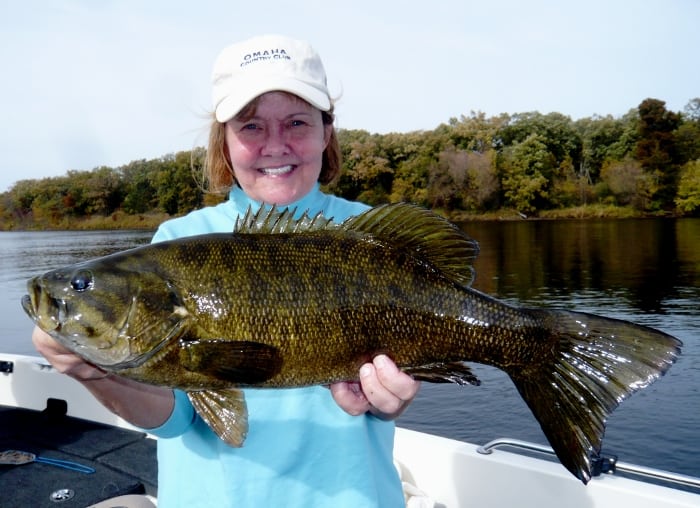 A smiling person wearing a white hat holds a large, freshly caught fish on a boat, enjoying fishing on a calm river near Alexandria, with trees in the background.