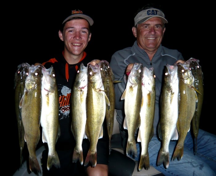 Two smiling men proudly hold up several freshly caught fish, displaying their successful summer walleye fishing haul against a dark background, likely at night. Both appear happy and content, dressed in casual outdoor clothing and caps.