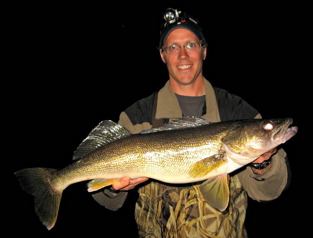 A person wearing glasses and a headlamp smiles while holding a large, shiny-scaled fish, posing against a dark background—just the kind of catch you’d expect to see in an Alexandria May Fishing Report.