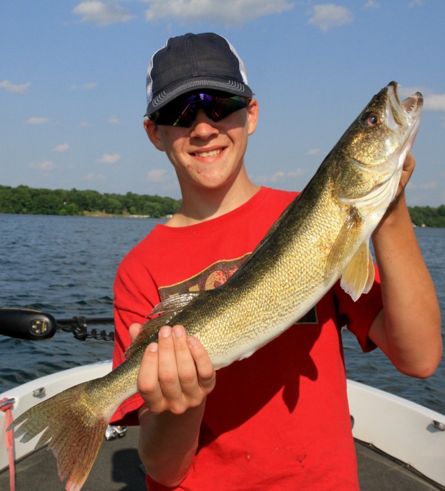 A smiling person in a red shirt and cap holds a large fish on a boat in a lake, showcasing summer fishing techniques against a backdrop of trees and blue sky.