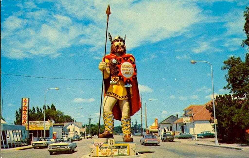 A large, colorful statue of a Viking warrior—known as "Big Ole" through the years—stands on a street median, surrounded by vintage cars, streetlights, and buildings under a blue sky with scattered clouds.