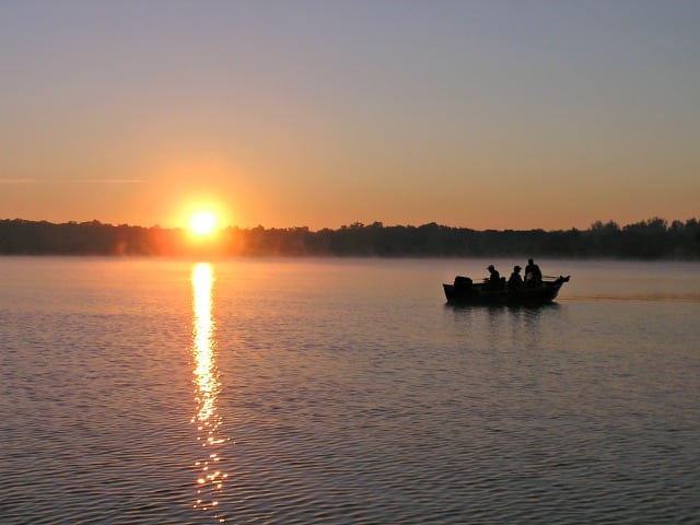 A small boat with three people is silhouetted against a calm lake during sunrise or sunset in Alexandria, MN, featured in 50 Photos Celebrating Alexandria, MN for Tourism Week. The sun casts a bright reflection as trees line the distant shore.
