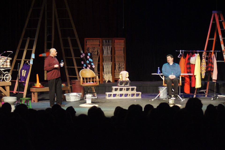Two actors perform on stage surrounded by props, costumes, and household items, with a seated audience watching. As they engage in dialogue, their scene humorously bids 2014 farewell and captures the festivity of New Year’s Celebrations.