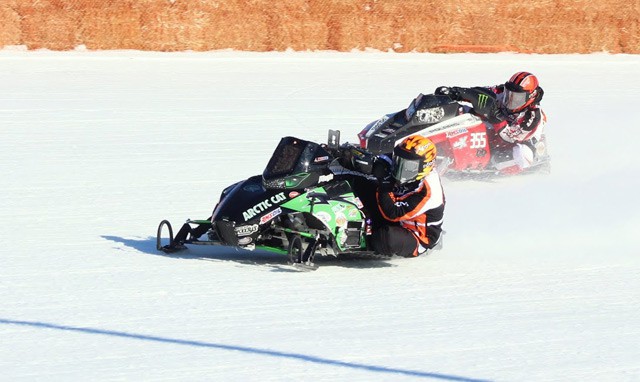 Two racers on snowmobiles speed around a bend on a snowy track, leaning into the turn as the spectacular tradition continues. The lead snowmobile is black and green, while the second is red and white. Both riders wear helmets and racing suits.