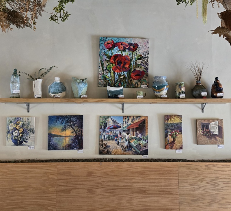 A gallery wall by the Alexandria Art Guild displays colorful paintings and ceramic vases on two shelves. Artworks include floral, landscape, and street scenes, with pottery and decorative items arranged beside them. Wooden panels cover the lower wall.