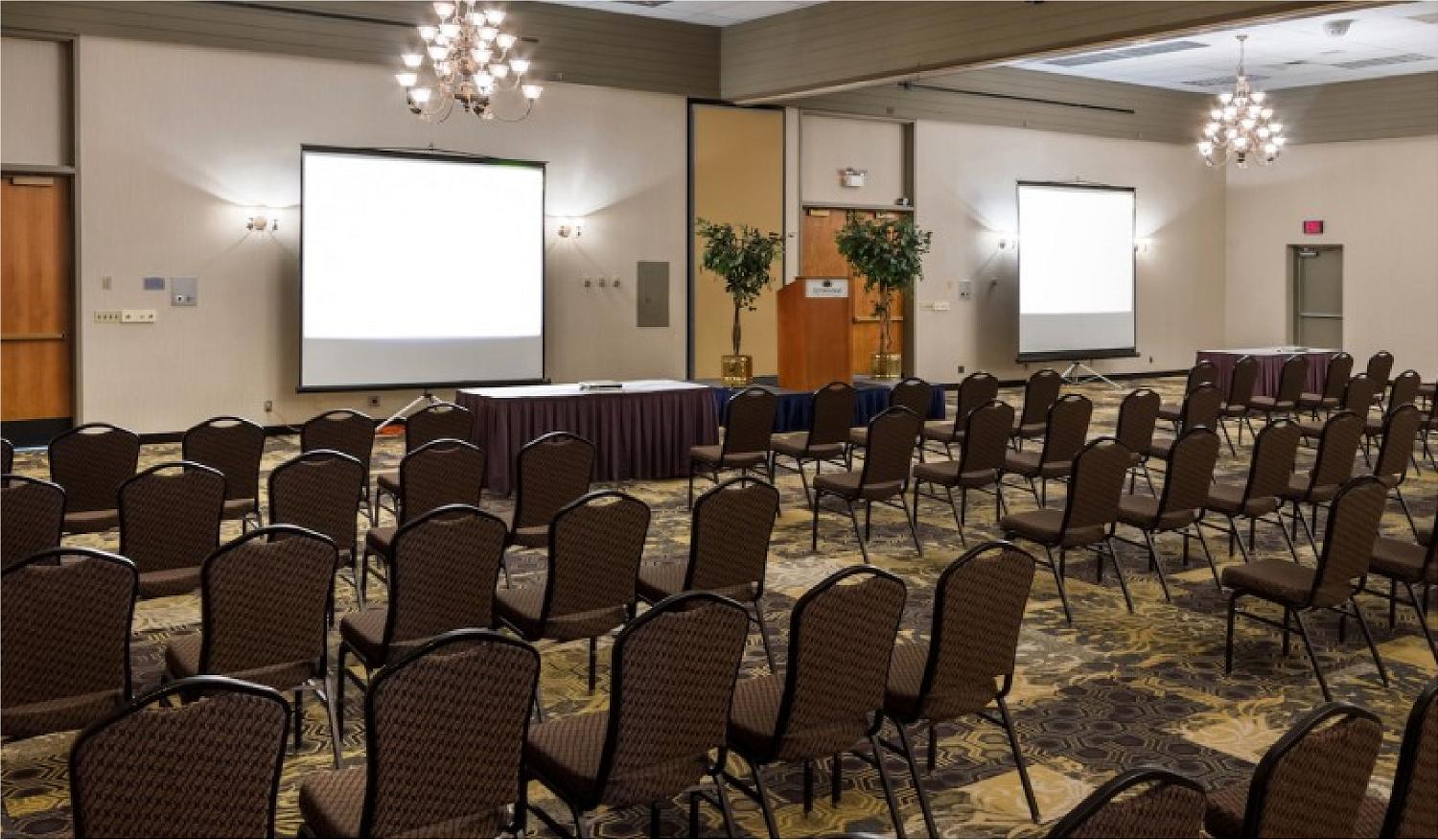 A conference room at Arrowwood Resort with rows of empty brown chairs facing two large projection screens, tables at the front, chandeliers overhead, decorative plants, and patterned carpeting.