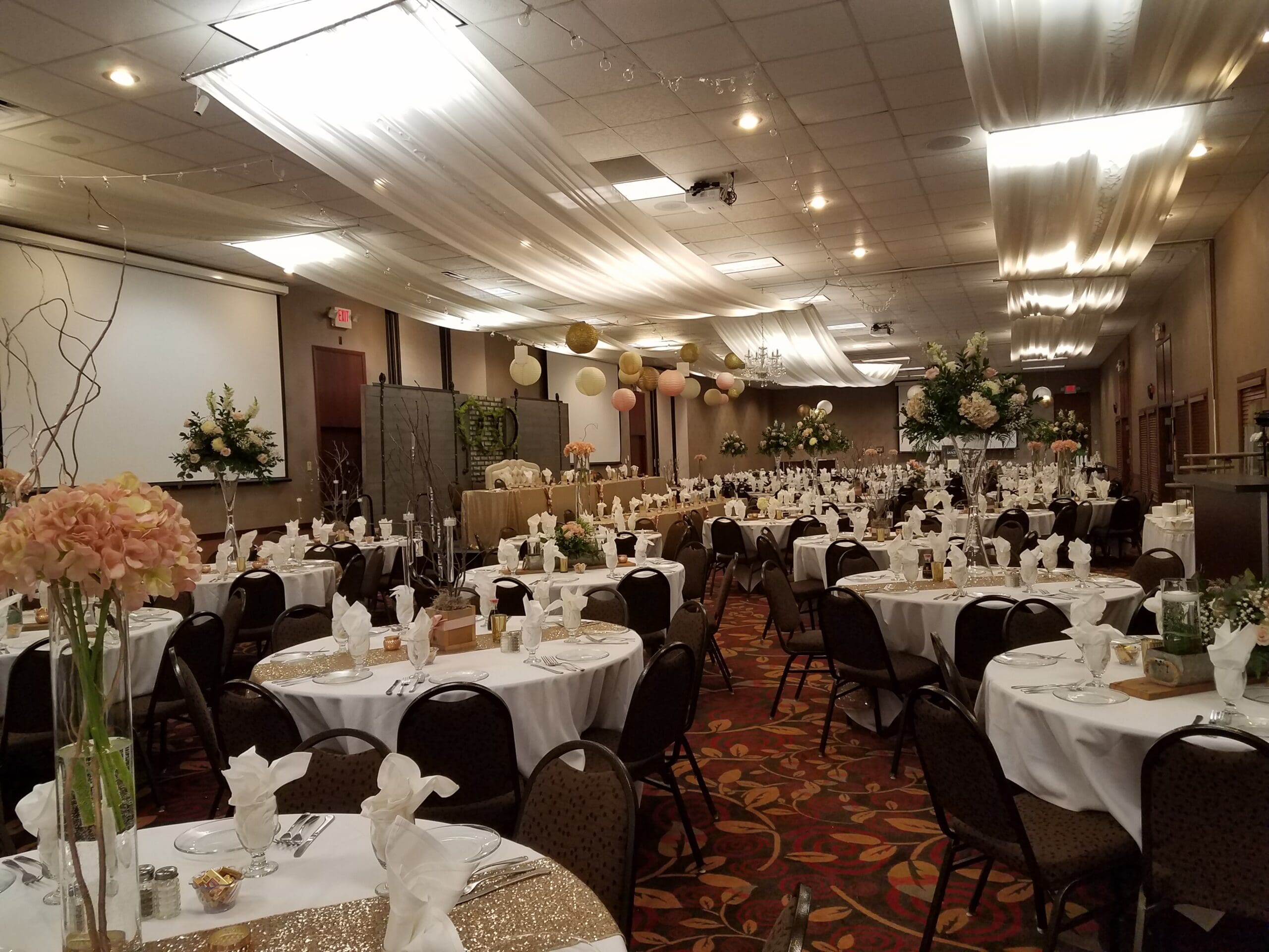 A large banquet hall at Holiday Inn decorated for an event with round tables covered in white tablecloths, folded napkins, floral centerpieces, draped white fabric, and hanging paper lanterns. The room is elegant and festive.