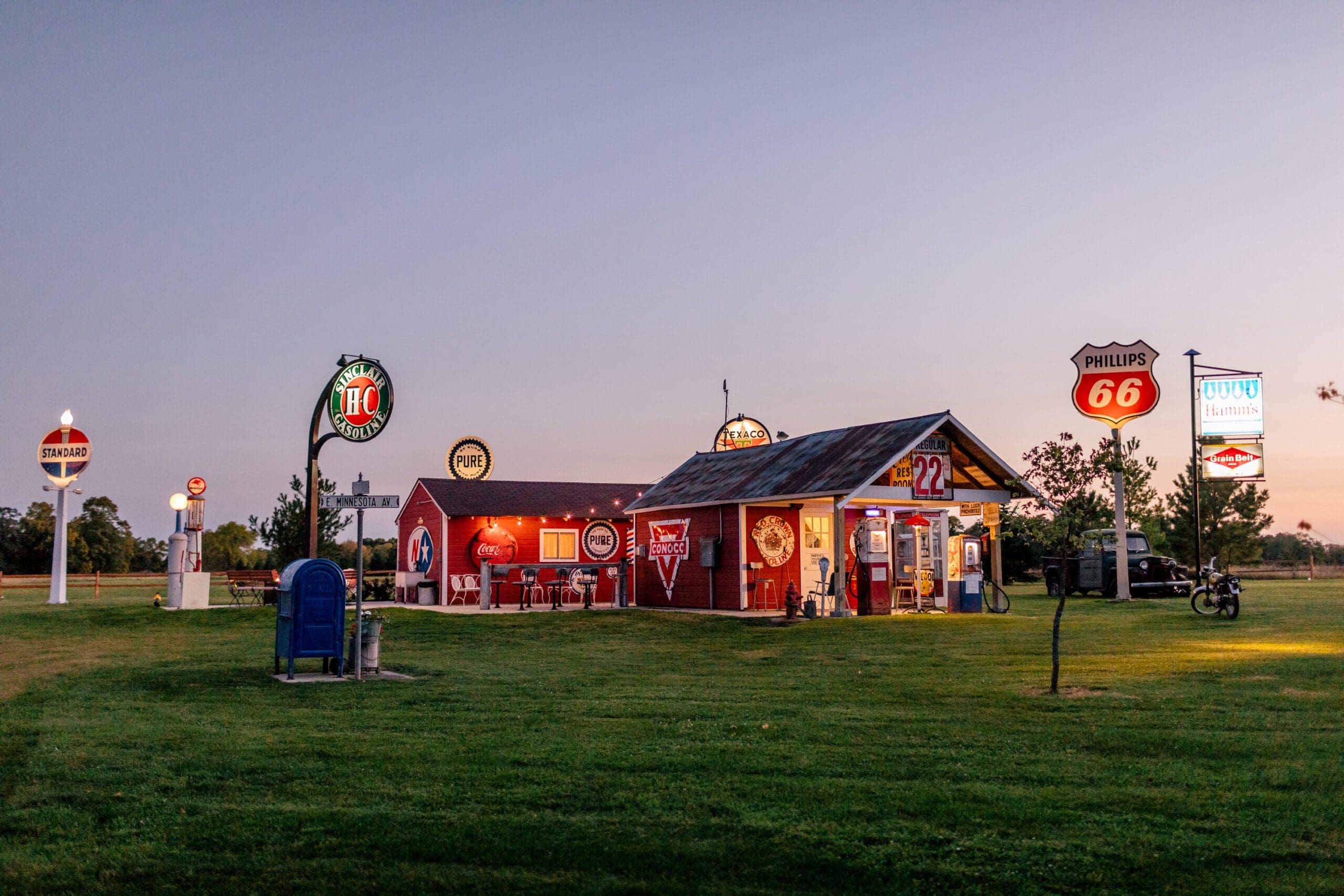 A roadside diner at Adler Ranch, with vintage gas station signs, outdoor seating, and glowing neon lights, sits on a grassy lawn at dusk. The clear sky reveals a few trees and a motorcycle in the background.