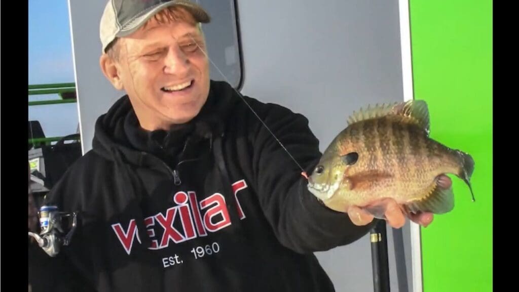 A smiling man named Mike in a cap and black "Vexilar" hoodie holds up a large sunfish by the mouth with a fishing rod in one hand, standing in front of a gray and green background.