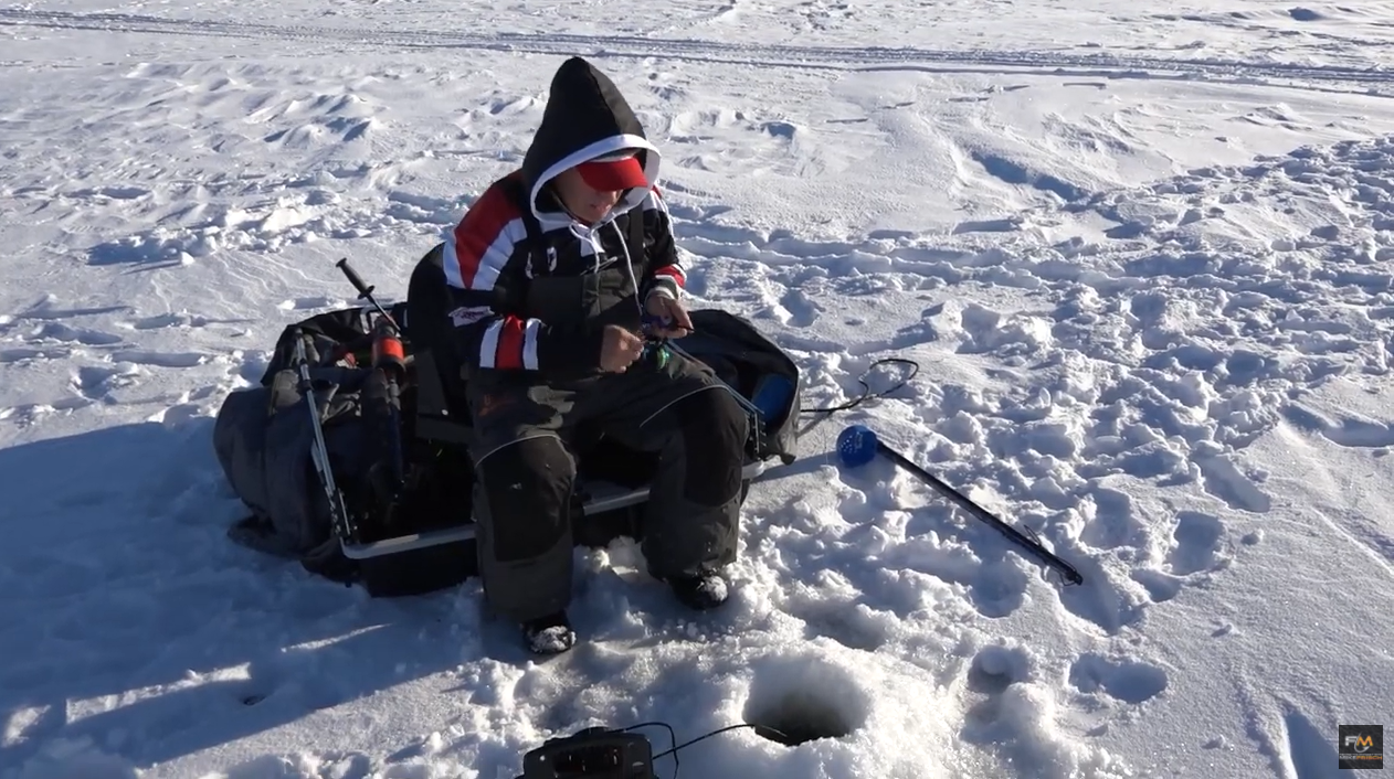A person in winter clothing sits on a sled, panfishing on a snowy frozen lake. They hold fishing gear near a hole in the ice, with an auger and other equipment nearby.
