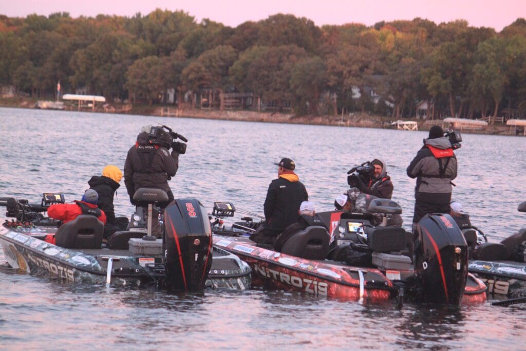 Several people in fishing boats, some with cameras, gather on a lake near a forested shoreline for a Major League Fishing event. The calm water and the soft light of sunrise or sunset create a picturesque scene.