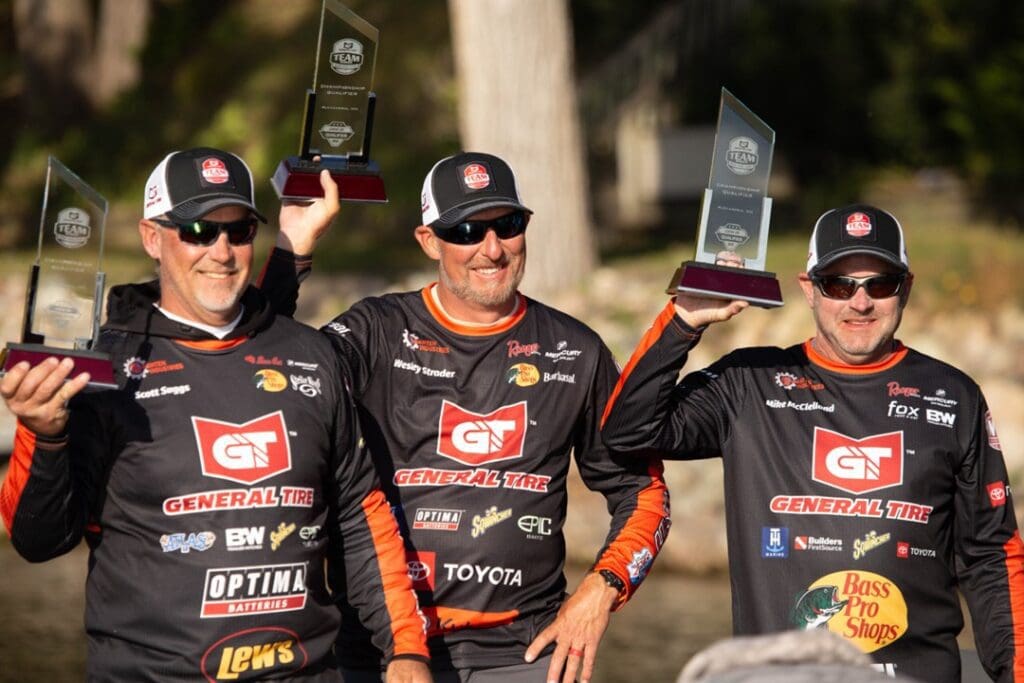 Three men wearing matching black and red General Tire fishing jerseys and hats smile while holding up clear glass trophies outdoors, with trees and sunlight in the background after a successful day of bass fishing.