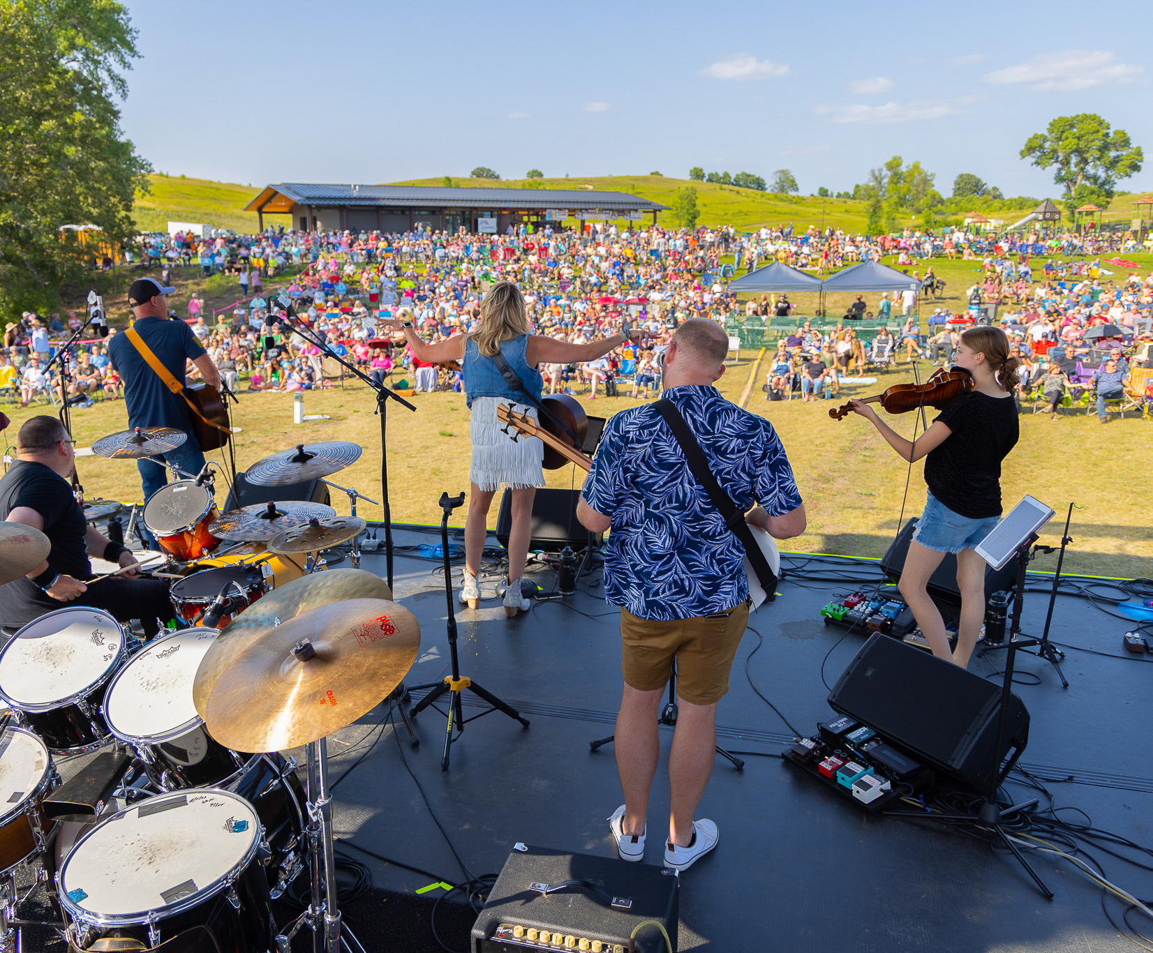 A band performs on an outdoor stage at Lake Brophy County Park, with a drummer, guitarists, and a violinist entertaining a large seated crowd on a sunny day in the grassy, open area.