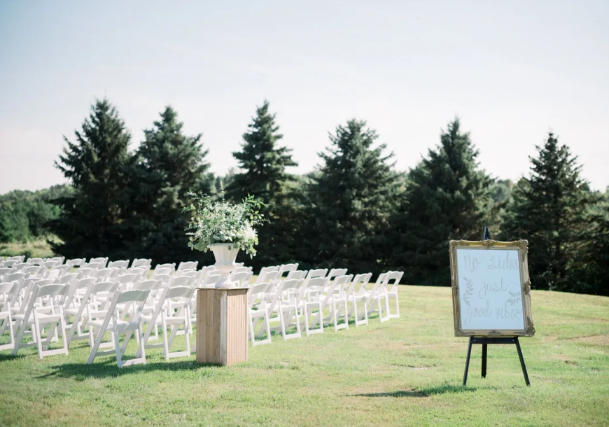 Rows of white chairs are set up outdoors on green grass at Rendezvous Farm, facing a large vase of greenery on a wooden stand. A framed sign on an easel stands nearby, with pine trees and a blue sky in the background.