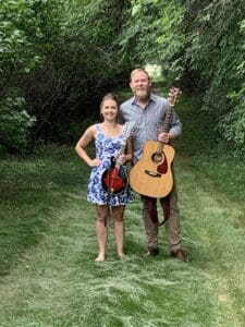 A woman in a floral dress with a violin and a man in a checked shirt holding an acoustic guitar stand together on a grassy path surrounded by green trees and bushes, ready to share live music perfect for an evening at 22 Northmen Brewing.