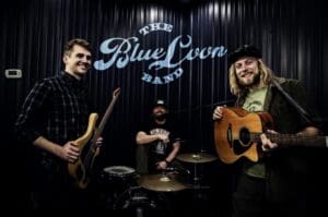 Three smiling musicians pose with instruments—an electric bass, an acoustic guitar, and a drum set—in front of a dark wall with "The Blue Loon Band" written on it at 22 Northmen Brewing, ready to bring live music to the crowd.
