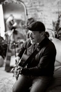 A man wearing a flat cap sits on a couch at 22 Northmen Brewing, playing an acoustic guitar and singing live music. In the background, a blurred reflection of another person with a guitar appears in a large ornate mirror. The photo is black and white.