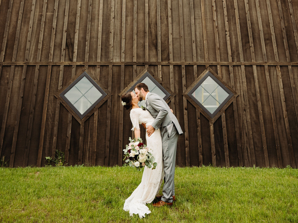 A bride and groom share a kiss in front of a rustic wooden barn with two diamond-shaped windows. The bride holds a bouquet of flowers while the groom gently dips her on green grass.