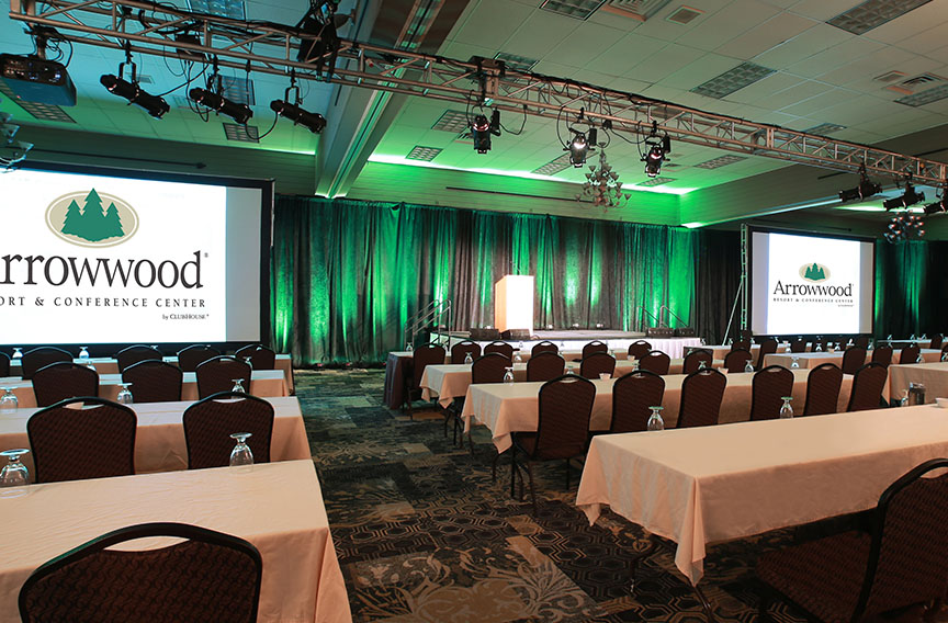 A conference room set up with rows of tables and chairs faces a stage with a podium, perfect for special events. Two large screens display "Arrowwood Resort & Conference Center," and the room is lit with green and white lighting.