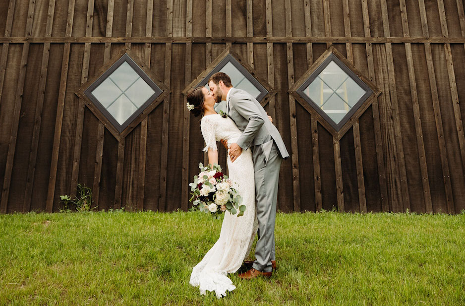 A bride and groom kiss in front of a rustic wooden barn with diamond-shaped windows during their Special Event. The bride holds a bouquet of flowers as the groom dips her on a green grassy lawn.