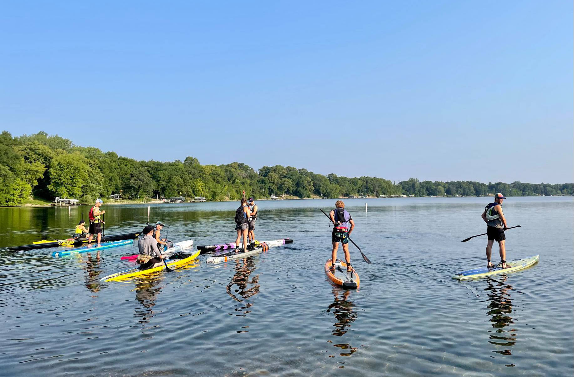 A group of people stand and kneel on paddleboards on a calm lake near the shore, surrounded by green trees under a clear blue sky.