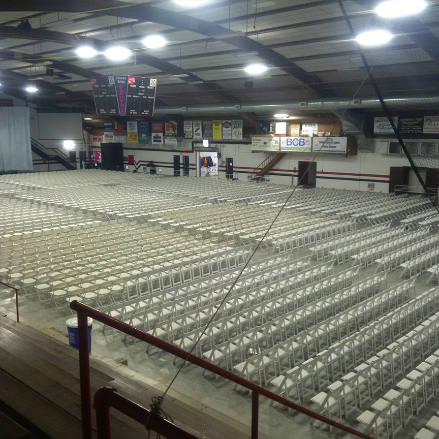 Rows of empty white folding chairs are arranged in an indoor sports arena, set up for conventions or meetings, all facing a small stage with audio equipment and a screen. Banners and scoreboards hang from the walls and ceiling.