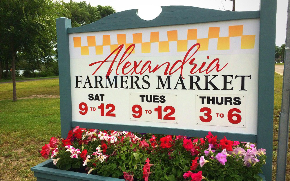 A sign for Alexandria Farmers Market, featured in the Summer Guide, lists hours: Saturday and Tuesday 9 to 12, Thursday 3 to 6. Red and pink flowers bloom in front of the sign, with grass and trees providing a natural backdrop.