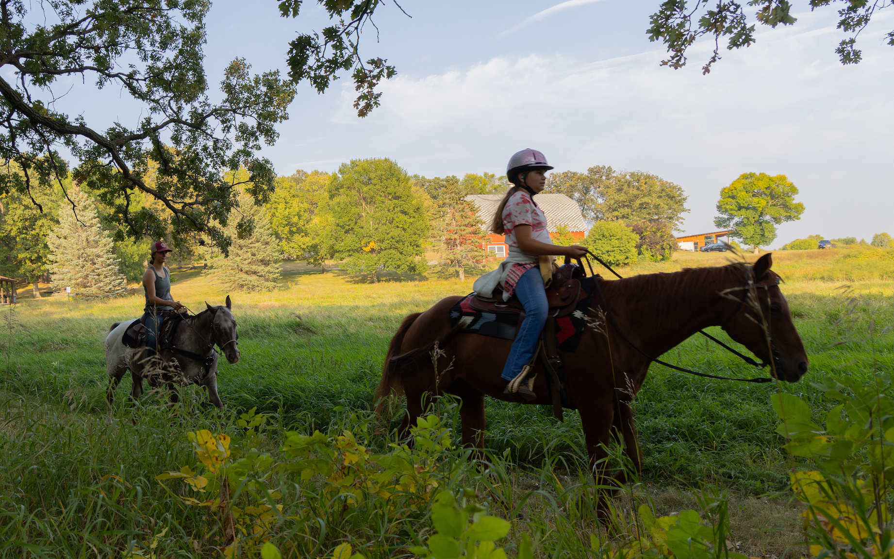Two people ride horses through a grassy, tree-filled area on a sunny day. Featured in our Summer Guide, the rider in front wears a helmet while the second follows behind. A farmhouse and trees can be seen in the background.