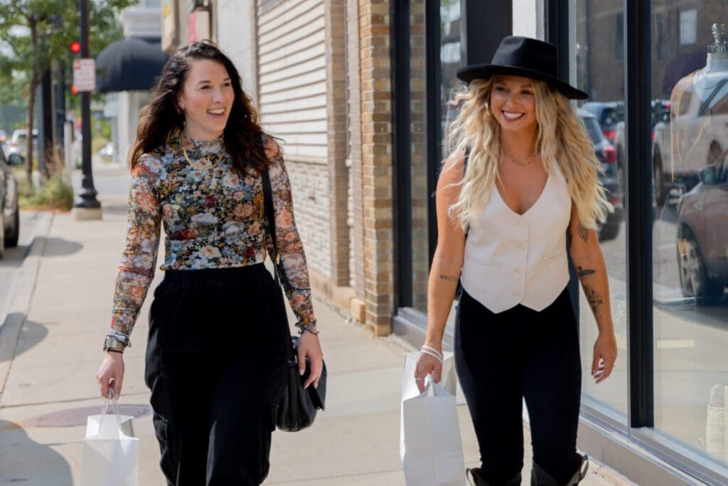 Two women smiling and walking down a city sidewalk, shopping bags in hand as they explore Alexandria MN. One wears a floral top and black pants, the other a white vest, black pants, and black hat. Shops and glass windows line the street.