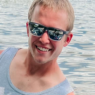 A smiling man wearing sunglasses and a light gray tank top stands near a body of water, reflections visible in his shades—a true fishing guide enjoying the outdoors.