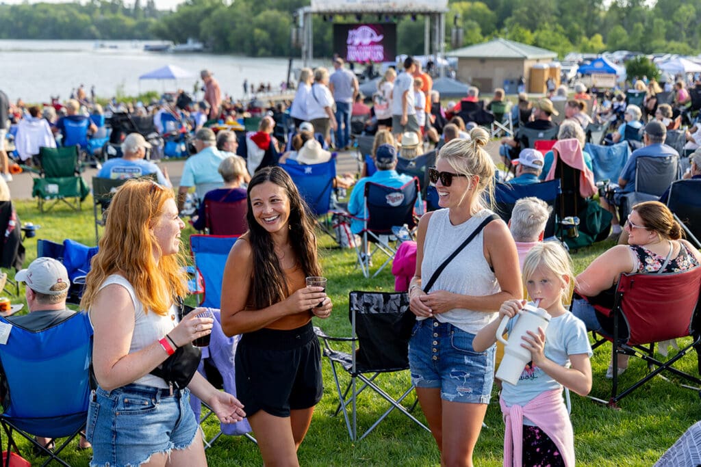 A group enjoys the Summer Sounds festival by Brophy Lake; three women and a child smile and chat in the foreground, while a crowd sits on lawn chairs facing the stage at this free summer concert.