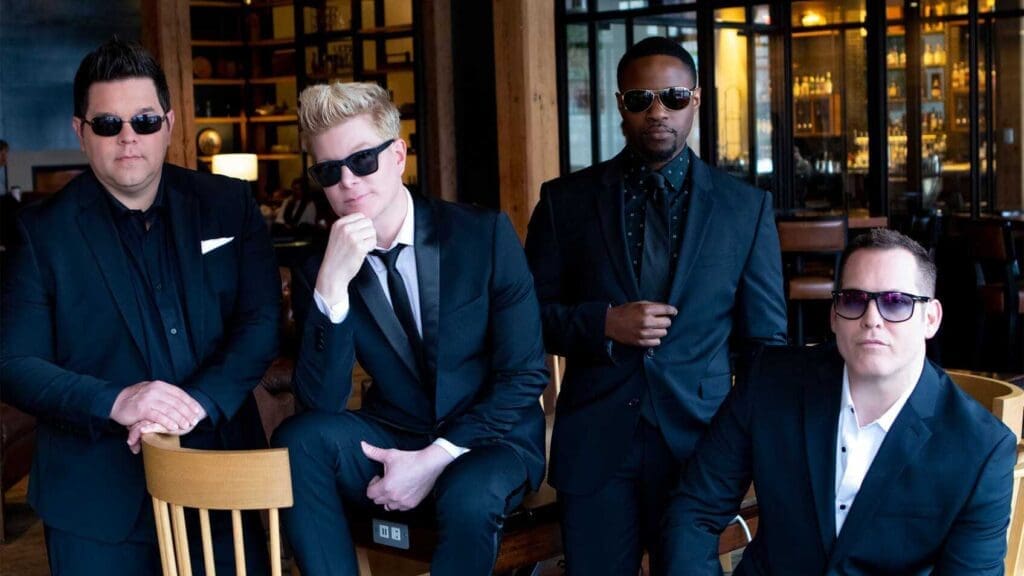 Four men in black suits and sunglasses pose confidently indoors, with two standing and two seated on chairs, in a stylish, modern setting with wooden elements and shelves in the background.