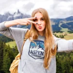 A young woman with long hair poses for a selfie outdoors at the Fall Pop-Up Shop, making a peace sign near her face. She wears a gray "Aspen Colorado" sweatshirt, with mountains and trees visible in the background.