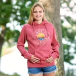 A young woman stands outdoors near Carlos Creek, smiling at the camera. She is wearing a pink “Lake Days” hoodie and denim shorts, her hands resting on her abdomen—ready to enjoy the Fall Pop-Up Shop by the water.