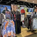 Clothing booth display featuring colorful hoodies, jackets, and shirts on racks, with large posters of smiling models wearing casual apparel in the background at the Carlos Creek Fall Pop-Up Shop expo.