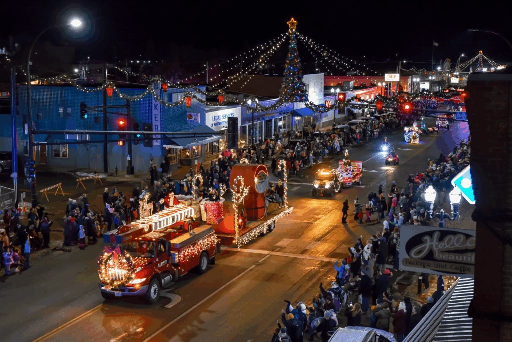 A nighttime holiday parade in Alexandria features brightly lit floats decorated with Christmas lights, a large crowd lining both sides of the street, and festive lights strung overhead, with a large Christmas tree in the background.