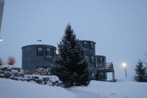 A snow-covered scene with two round, silo-like buildings, a large evergreen tree in the center, and a snowy path leading up to the structures. A lamp post glows faintly in the evening light—an inviting place to beat the winter blues.