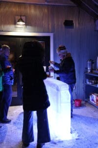 A person in a winter coat stands at an illuminated ice bar, chasing away the Winter Blues while another person serves drinks; two more people wait nearby in a dimly lit, industrial-style room.