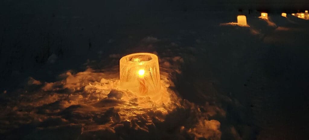 A candle glows inside a clear ice lantern placed on snow at night at Lake Carlos State Park, with more ice lanterns lining a snowy path in the background, creating a warm, peaceful atmosphere during the Candlelight Event.